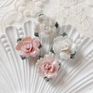 May include: Four iridescent flower-shaped embellishments in shades of pink and white, with green leaf accents. The floral decorations are arranged on a white, scalloped surface, creating a delicate and elegant aesthetic.
