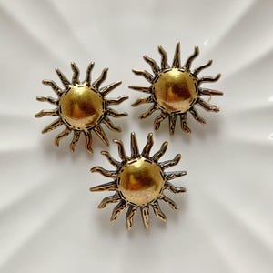 Puede incluir: Tres botones de metal de color dorado en forma de sol con un diseño en relieve texturizado.