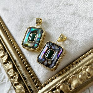Può includere: Due ciondoli dorati di forma quadrata. Ogni ciondolo presenta una pietra preziosa verde circondata da madreperla di abalone iridescente. I ciondoli sono su una superficie bianca.