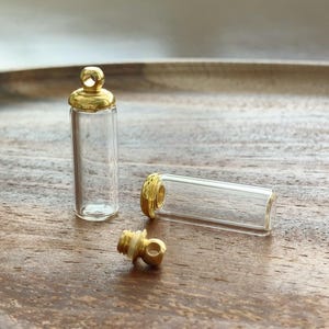 May include: Two small, clear glass vials with gold-coloured metal screw-top caps. One vial stands upright, while the other lies horizontally. The detached gold cap sits in the foreground, revealing a small loop at the top for hanging or attachment.
