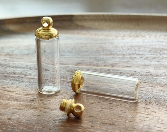 以下が含まれることがあります： 金色の金属製スクリューキャップが付いた、2つの小さな透明なガラス瓶。1つは直立しており、もう1つは横向きに置かれています。手前には取り外された金色のキャップがあり、吊り下げや取り付け用の小さなループが見えます。