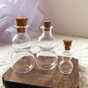 Puede incluir: Tres botellas de vidrio transparente con tapones de corcho, dispuestas sobre una bandeja de madera. Las botellas tienen una forma única de doble bulbo. Los corchos son de color marrón claro. La bandeja es de color marrón oscuro.