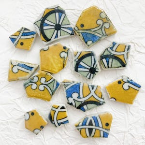 Puede incluir: Colección de baldosas de cerámica rotas con un diseño amarillo, azul y blanco. Las baldosas tienen un acabado brillante y presentan patrones geométricos y florales. Las piezas están dispersas sobre una superficie blanca texturizada.