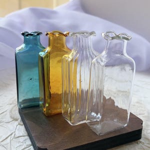 May include: Four small, square glass vases in shades of blue, yellow, and clear, arranged on a dark wooden base. Each vase has a ruffled top. The vases are displayed against a soft, white background.