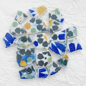May include: A collection of irregularly shaped ceramic tiles with geometric patterns. The tiles feature a white base with designs in blue, green, yellow, and gray. The tiles are arranged on a white, textured surface.