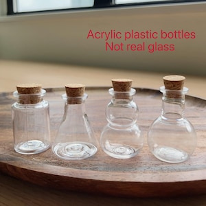 May include: Four clear acrylic plastic bottles with cork stoppers in various shapes including cylindrical, conical, double-bulb, and round. The text Acrylic plastic bottles Not real glass is visible above the bottles.