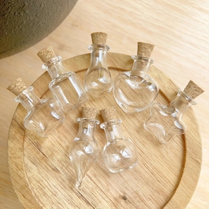 May include: A collection of small, clear glass bottles with cork stoppers. The bottles are various shapes, including a heart, a star, and a teardrop. They are arranged on a round, wooden tray.