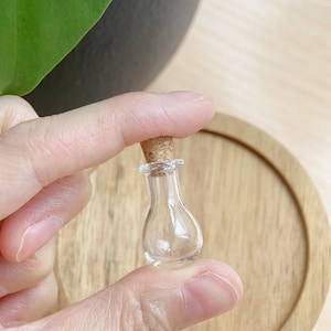 Puede incluir: Una pequeña botella de vidrio transparente con un tapón de corcho. La botella tiene un cuerpo bulboso y un cuello estrecho. El corcho es de color marrón claro. La botella se sostiene entre dos dedos sobre un fondo de madera.