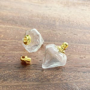 Könnte beinhalten: Zwei klare, rautenförmige Glasfläschchen mit goldfarbenen Schraubverschlüssen aus Metall. Ein Fläschchen ist mit dem Verschluss montiert, während das andere mit seinem kleinen, abnehmbaren Gewindestopfen aus Goldmetall daneben liegt.