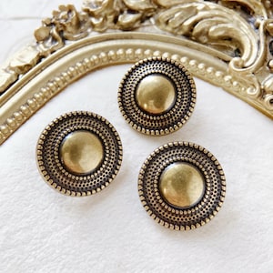 Op de afbeelding: Drie antieke messingkleurige kastknoppen. Elke knop heeft een centrale, iets verhoogde cirkel omgeven door een ring van kleine, gelijkmatig verdeelde stippen. De knoppen zijn rond en hebben een vintage esthetiek.