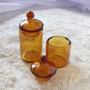 May include: Three amber-colored glass containers. One is a tall, cylindrical jar with a lid and a round knob. Another is a shorter, open cylinder. The third is a small, lidded jar with a rounded base and a knob on top. All are made of transparent glass.