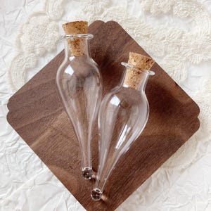 Puede incluir: Dos botellas de vidrio transparente con tapones de corcho sobre una bandeja de madera marrón oscuro. Las botellas tienen forma de lágrima con una base pequeña y redonda. Los corchos son de color marrón claro. La bandeja tiene un borde decorativo.