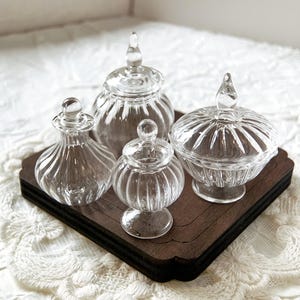 May include: Four clear glass jars with lids, some with a ribbed design, on a dark brown wooden tray. The jars are empty and have a vintage look.