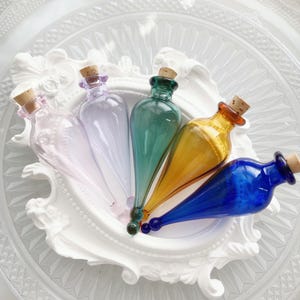 May include: Five teardrop-shaped glass bottles with cork stoppers, arranged in a fan pattern. The bottles are translucent in shades of light pink, pale purple, emerald green, amber, and deep blue, each featuring a small spherical base tip.