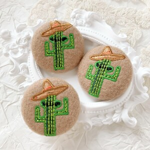May include: Three circular, tan felt brooches featuring embroidered cactus designs. Each cactus wears a small, brown sombrero with a red trim. The cacti are green with black details. The brooches are displayed on a white, decorative plate.