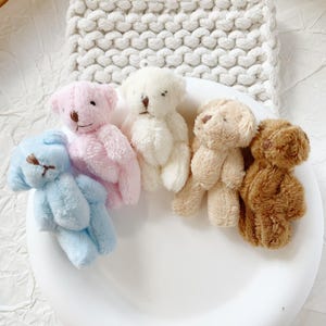May include: Five miniature teddy bears in pastel shades of blue, pink, white, beige, and brown. Each bear has a stitched nose and button eyes. They are arranged on a white surface, with a white knitted background.