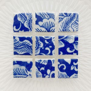 May include: Nine square ceramic tiles with a blue and white floral pattern. The tiles are arranged in a 3x3 grid on a clear, textured surface. Each tile features a unique design of leaves and flowers.
