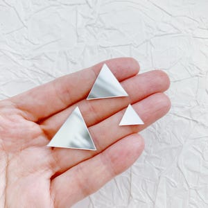 Könnte beinhalten: Vier kleine, reflektierende, silberfarbene Dreiecke unterschiedlicher Größe. Sie werden in einer Hand vor einem weißen, strukturierten Hintergrund gehalten. Die Dreiecke scheinen aus glänzendem Metall gefertigt zu sein.