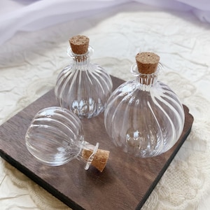 Può includere: Tre bottiglie di vetro trasparente con tappi di sughero su un vassoio di legno scuro. Le bottiglie hanno un design sferico a coste. I tappi sono di colore marrone chiaro. Il vassoio ha un bordo smerlato.