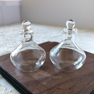 Puede incluir: Dos frascos de perfume de vidrio transparente con cuerpos redondeados y pequeños tapones redondos. Los frascos se exhiben sobre una superficie de madera oscura. El vidrio refleja la luz, creando reflejos y sombras.