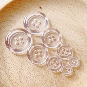 May include: A set of eight clear plastic buttons of varying sizes. The buttons have four holes each.