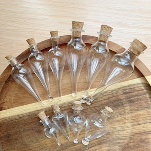 May include: A selection of clear glass bottles with cork stoppers, displayed on a wooden tray. The bottles are teardrop-shaped and come in different sizes. The tray is made of wood and has a natural grain.