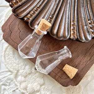 Pode incluir: Duas pequenas garrafas de vidro transparente com rolhas de cortiça. As garrafas têm a forma de um diamante e um fundo arredondado. As garrafas estão sobre uma superfície de madeira.
