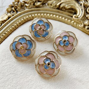 Puede incluir: Cuatro pendientes con forma de flor con borde dorado. Cada pendiente presenta un diseño floral en azul claro, rosa y blanco. Los pendientes están dispuestos sobre una superficie blanca. Los pendientes miden unos 2 cm de diámetro.