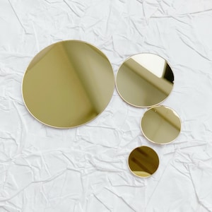May include: Four gold-coloured, reflective, circular mirrors of varying sizes are arranged on a white, textured surface. The largest mirror is at the top left, with the smallest at the bottom right. The mirrors have a shiny, metallic appearance.