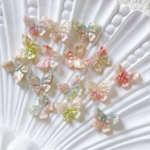 May include: A selection of iridescent butterfly embellishments in pastel shades of pink, yellow, and blue. The butterflies are displayed on a white, textured surface, highlighting their delicate details and shimmering appearance.