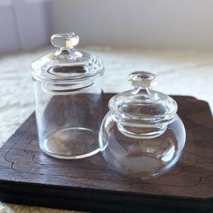 Könnte beinhalten: Zwei transparente Glasbehälter mit Deckeln. Ein Behälter ist zylindrisch, der andere rund. Beide Behälter haben einen kleinen, transparenten Knopf auf dem Deckel. Die Gläser stehen auf einem dunkelbraunen Holztablett.