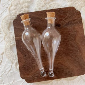 Puede incluir: Dos botellas de vidrio transparente con tapones de corcho. Las botellas tienen una forma de lágrima única con una base bulbosa y una pequeña bola de vidrio redonda en la parte inferior. Se exhiben sobre una superficie de madera oscura.