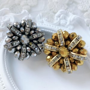 Puede incluir: Dos broches decorativos con forma floral. Un broche es plateado con detalles de cristal transparente, y el otro es dorado con detalles de cristal transparente. Ambos broches tienen una gema central facetada, sobre una superficie blanca.