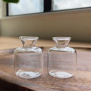 Op de afbeelding: Twee kleine, heldere glazen flesjes met een afgerond lichaam en een uitlopende, open hals. De flesjes zijn transparant en hebben een gladde, cilindrische vorm met een brede basis en een smalle opening aan de bovenkant voor kleine hoeveelheden.