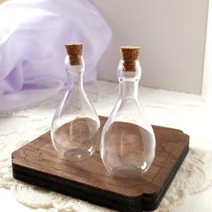 May include: Two clear glass bottles with cork stoppers sit on a dark brown wooden tray. The bottles have a teardrop shape and are empty. The corks are a light brown color. The tray has a decorative edge.