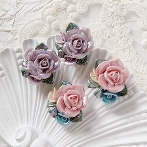Könnte beinhalten: Vier keramische Rosenblüten-Dekorationen in Rosa- und Lavendeltönen mit schillernden grünen Blättern. Die Rosen sind auf einer weißen, strukturierten Oberfläche angeordnet.