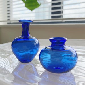 May include: Two small cobalt blue glass vases. One vase features a tall, slender neck with a rounded body, while the other is shorter with a wider, squat, bulbous shape. Both vessels have smooth, translucent surfaces and circular openings at the top.