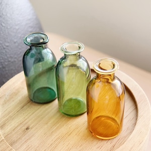 May include: Three small glass bottles in shades of green and amber sit on a wooden tray. The bottles have a rounded shape with narrow necks. The colors of the bottles vary from a deep emerald to a light amber, creating a visually appealing arrangement.