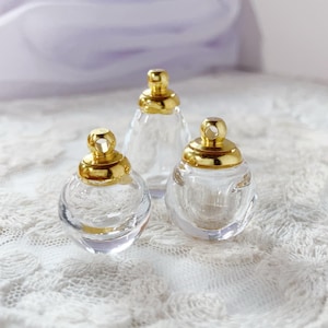 Peut inclure: Trois flacons de parfum en verre transparent avec des bouchons dorés. Les flacons ont des formes différentes : ronde, en forme de goutte et triangulaire. Les flacons sont présentés sur une surface en dentelle blanche.