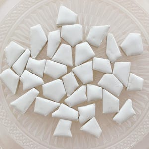 Op de afbeelding: Een verzameling kleine, onregelmatige, witte keramische mozaïektegels met gladde, glanzende oppervlakken. Elk stuk heeft een unieke, veelzijdige geometrische vorm en ligt verspreid over een doorzichtig, gestructureerd glazen bord.