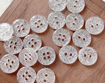 May include: A collection of 17 silver glitter buttons with four holes each. The buttons are arranged in a random pattern on a wooden surface.