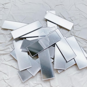 May include: A collection of small, rectangular, silver-colored metal pieces scattered on a crumpled white surface. The pieces have a reflective, mirror-like finish, and some appear to have a protective film.