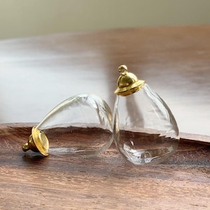 Puede incluir: Dos colgantes de cristal transparente en forma de lágrima con tapas de metal dorado. Un colgante está tumbado de lado, mientras que el otro está en posición vertical, con una pequeña anilla de metal en la parte superior para colgar.