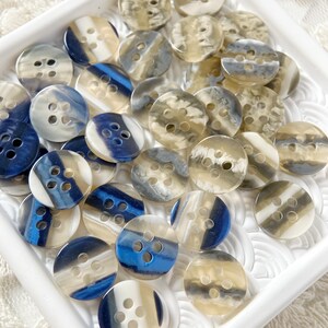 Op de afbeelding: Een verzameling ronde, witte en blauwe knopen met elk vier gaten. De knopen hebben een marmereffect met blauwe en witte swirls.