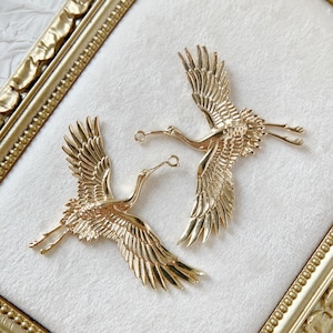 Könnte beinhalten: Zwei goldfarbene Metall-Kranich-Anhänger mit filigranen Details. Die Kraniche sind in entgegengesetzte Richtungen gerichtet und haben oben eine Schlaufe zum Anbringen an Schmuck oder anderen Bastelarbeiten.