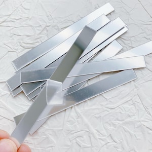 May include: A collection of thin, rectangular, silver metal strips. The strips are arranged in a slightly overlapping pattern, with some appearing to be bent. The background is a textured, white surface.