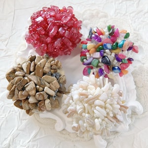 May include: Four decorative napkin rings, each with a unique design. One is made of red crystals, another of colorful stones, a third of beige pebbles, and the last of white and beige shell fragments.