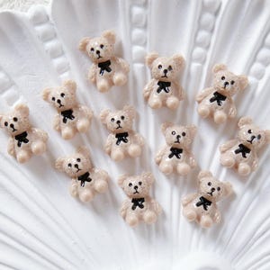 Könnte beinhalten: Eine Ansammlung kleiner, hellbrauner Teddybär-Figuren mit schwarzen Fliegen. Die Bären haben schwarze Augen und sind auf einer weißen, muschelförmigen Oberfläche angeordnet. Diese Miniatur-Bären sind wahrscheinlich zum Basteln oder Dekorieren.