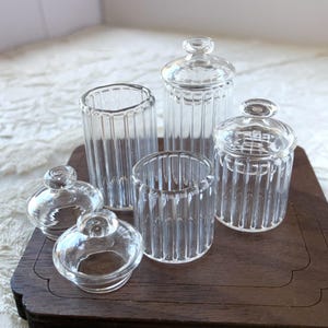 Op de afbeelding: Een verzameling heldere glazen potten en deksels met verticale ribbels. De set bevat potten in verschillende maten, sommige met deksels, gerangschikt op een donkerhouten dienblad. De transparante potten reflecteren het licht.