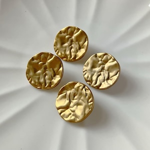 Könnte beinhalten: Vier goldfarbene, runde Ohrringe mit gehämmerten Strukturen liegen auf einer weißen Oberfläche. Die Ohrringe haben eine leicht unregelmäßige Form mit mattem Finish. Die Ohrringe sind wahrscheinlich aus Metall und für den täglichen Gebrauch geeignet.